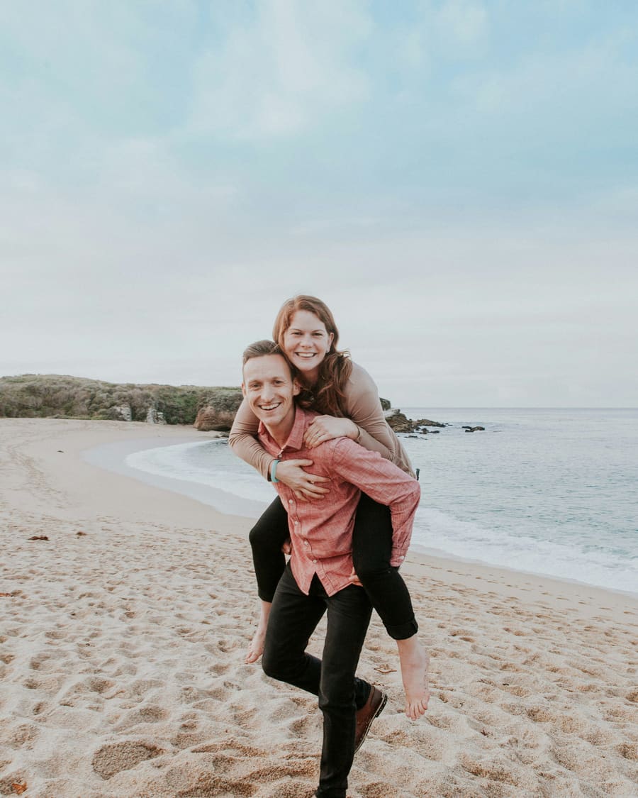 A woman riding piggyback on her companion, a man in a pink shirt, both smiling on a sandy beach under a pale sky.