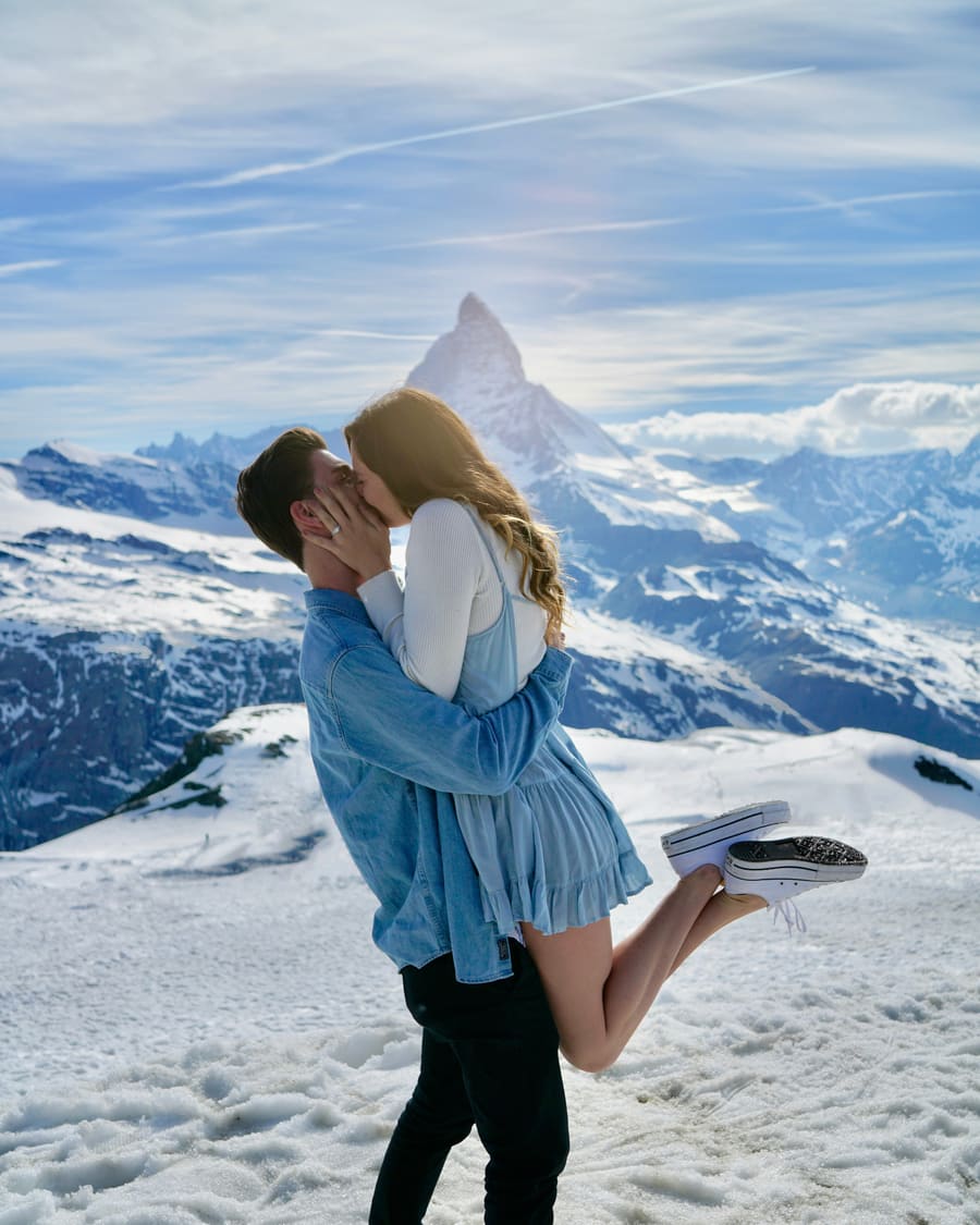 Eine Frau im blauen Kleid wird von ihrem Begleiter, einem Mann in Jeansjacke, hochgehoben, während sie sich im tiefen Schnee unter einem spitzen Alpengipfel küssen.