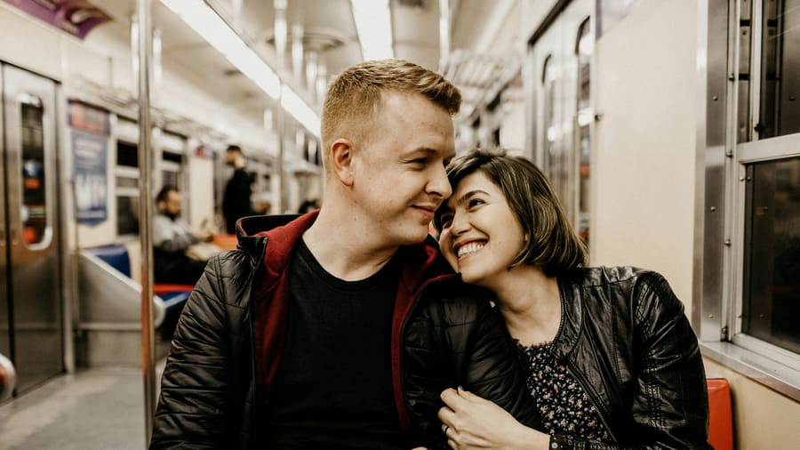 Eine Frau mit kurzen dunklen Haaren neigt lächelnd den Kopf zu ihrem Begleiter, einem Mann in dunkler Jacke, auf einem Sitz in einem U-Bahn-Wagen.