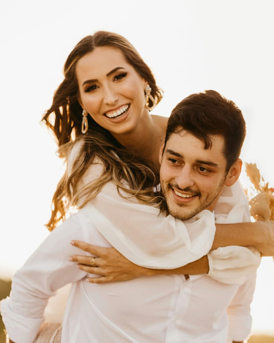 A woman in a white blouse and drop earrings laughing as she rides piggyback on her companion, a dark-haired man in a white shirt, in warm backlight.