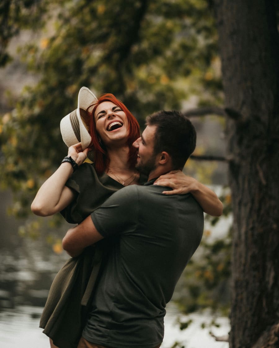 A red-haired woman holding a straw hat and laughing as her companion, a man in a dark T-shirt, lifts her beside a lake under trees.