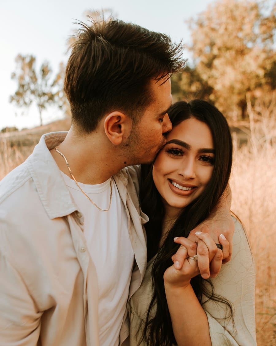A woman with long dark hair smiling at the camera as her companion, a man in a cream shirt, kisses her cheek in a field of dry grass at golden hour.
