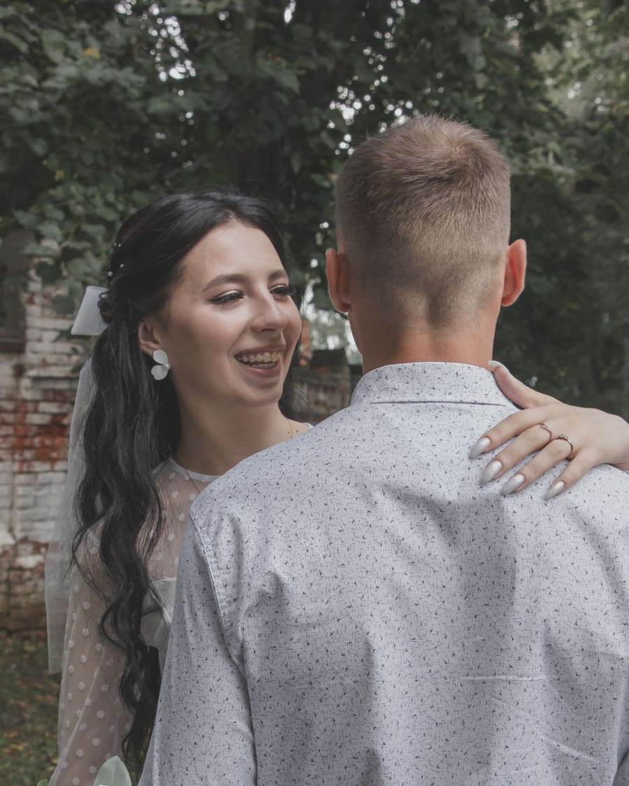 A woman in a white dress and veil, a bow in her hair, smiling at her companion, a man in a patterned shirt seen from behind, her hand on his shoulder, beside an old brick wall under trees.