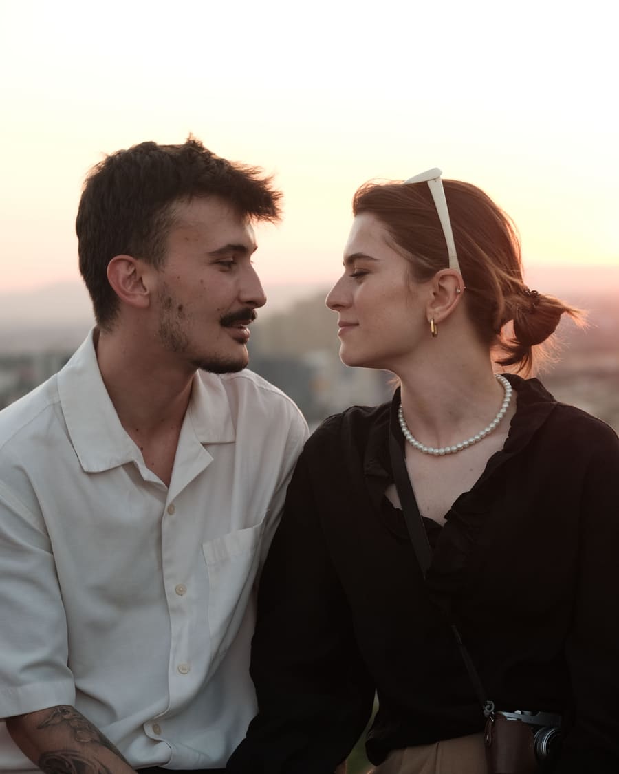 A woman wearing a pearl necklace and her companion, a man in a white shirt, smiling at each other on a rooftop above the city at sunset.