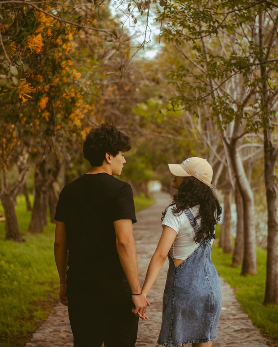 A woman in a denim pinafore and a cap walking hand in hand with her companion, a man in a black T-shirt, down a tree-lined stone path.