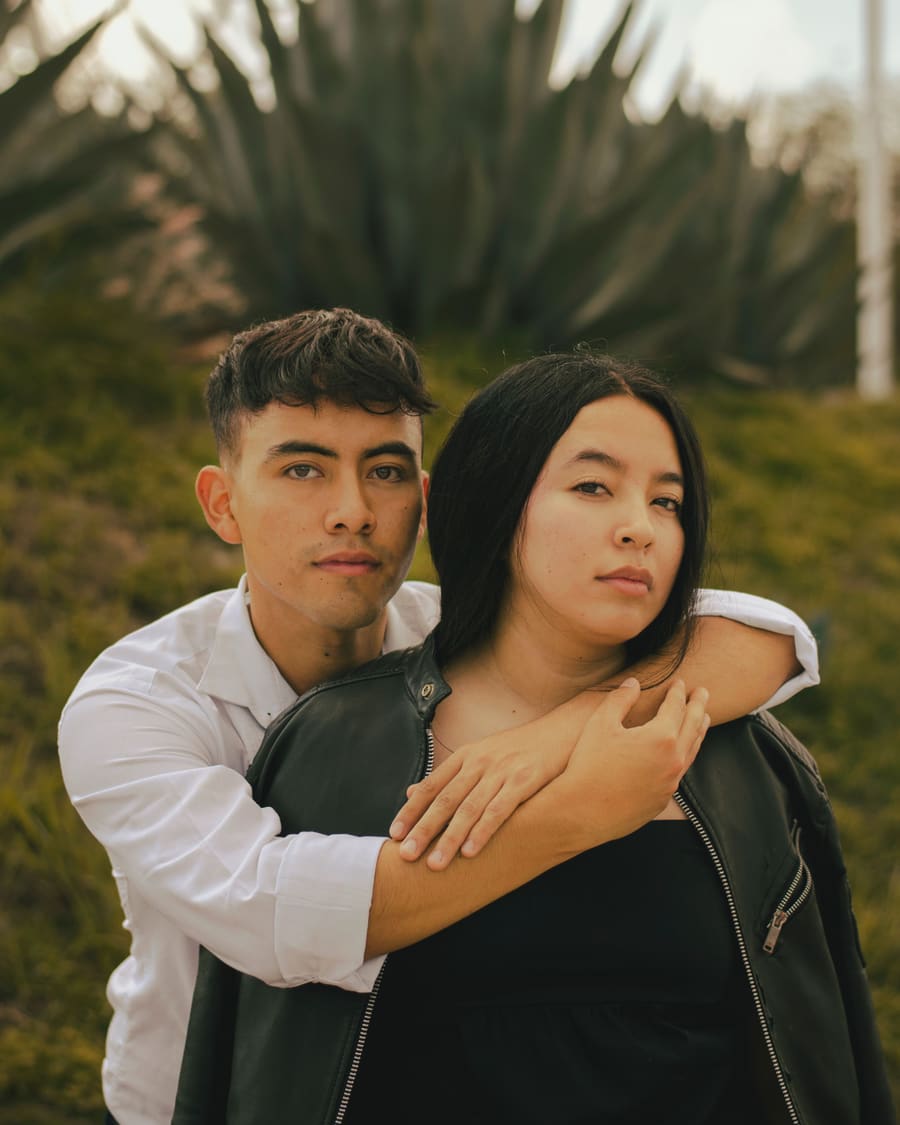 A woman in a leather jacket looking at the camera as her companion, a man in a white shirt, wraps his arms around her from behind in front of a large agave.
