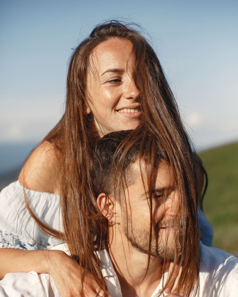A freckled woman hugging her companion from behind, her long hair blowing across his face, as they smile on a sunny hillside.
