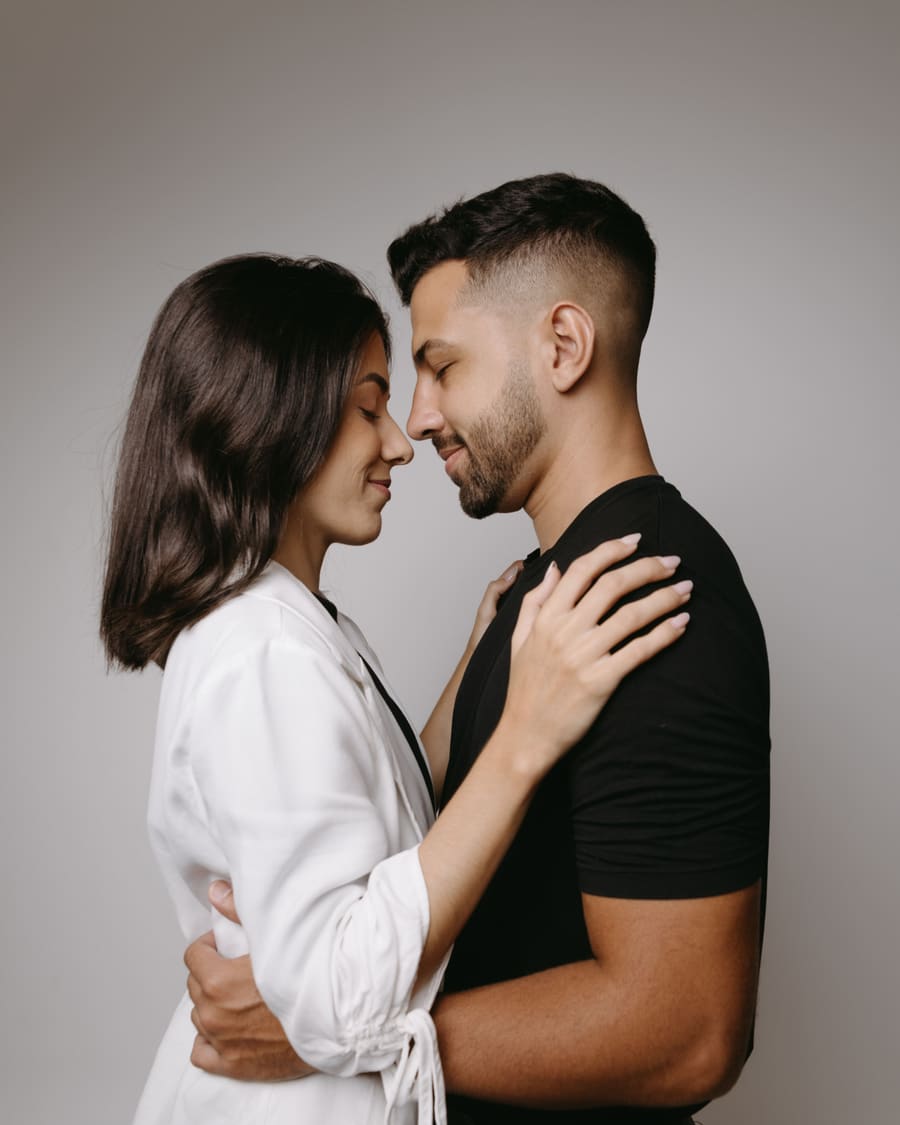 A woman in a white shirt with her hand on the shoulder of her companion, a man in a black T-shirt, their faces close, against a plain grey wall.