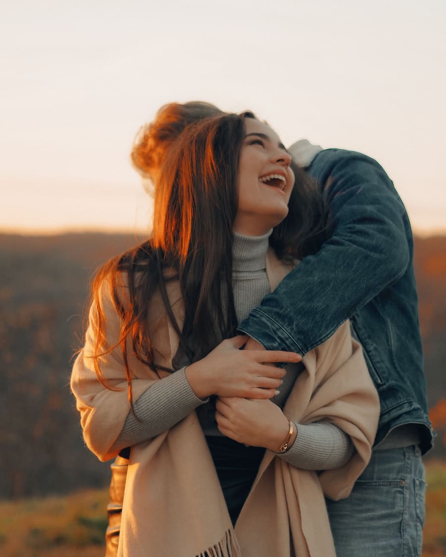 A laughing woman in a beige scarf hugged from behind by her companion, a man in a denim jacket, on a hilltop at sunset.