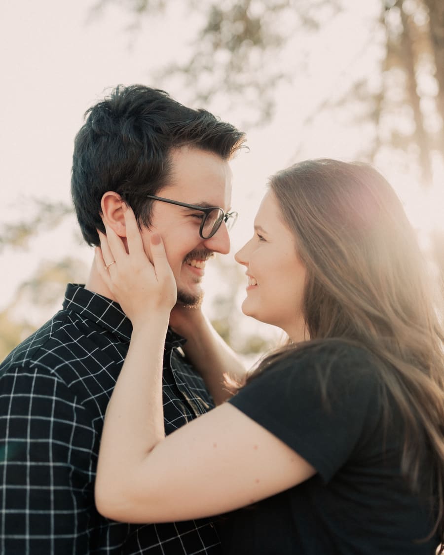 A woman touching the face of her companion, a man in glasses and a checked shirt, both smiling in soft afternoon light.