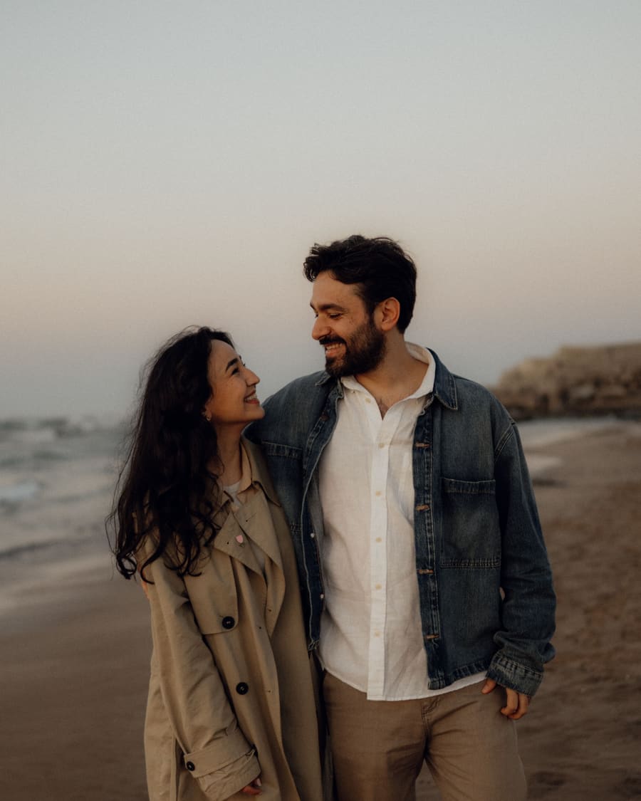 Eine Frau im beigen Trenchcoat und ihr Begleiter, ein Mann in Jeansjacke, lachen gemeinsam in der Abenddämmerung an einem Strand.