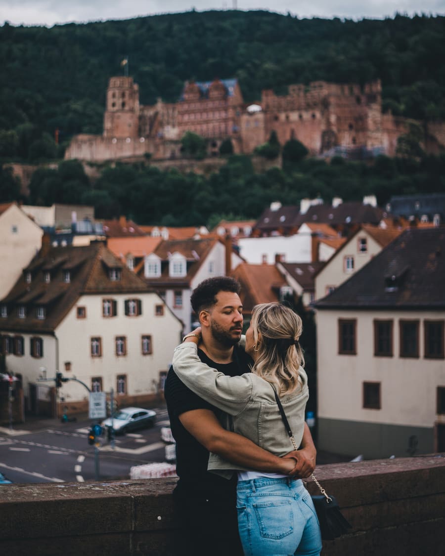 Eine Frau in heller Jacke umarmt ihren Begleiter, einen Mann in Schwarz, an einer Steinmauer über roten Dächern unterhalb des Heidelberger Schlosses.