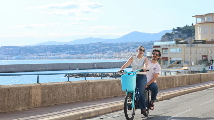 Eine Frau und ihr Begleiter fahren gemeinsam auf einem Fahrrad über eine Uferpromenade an einer blauen Bucht.