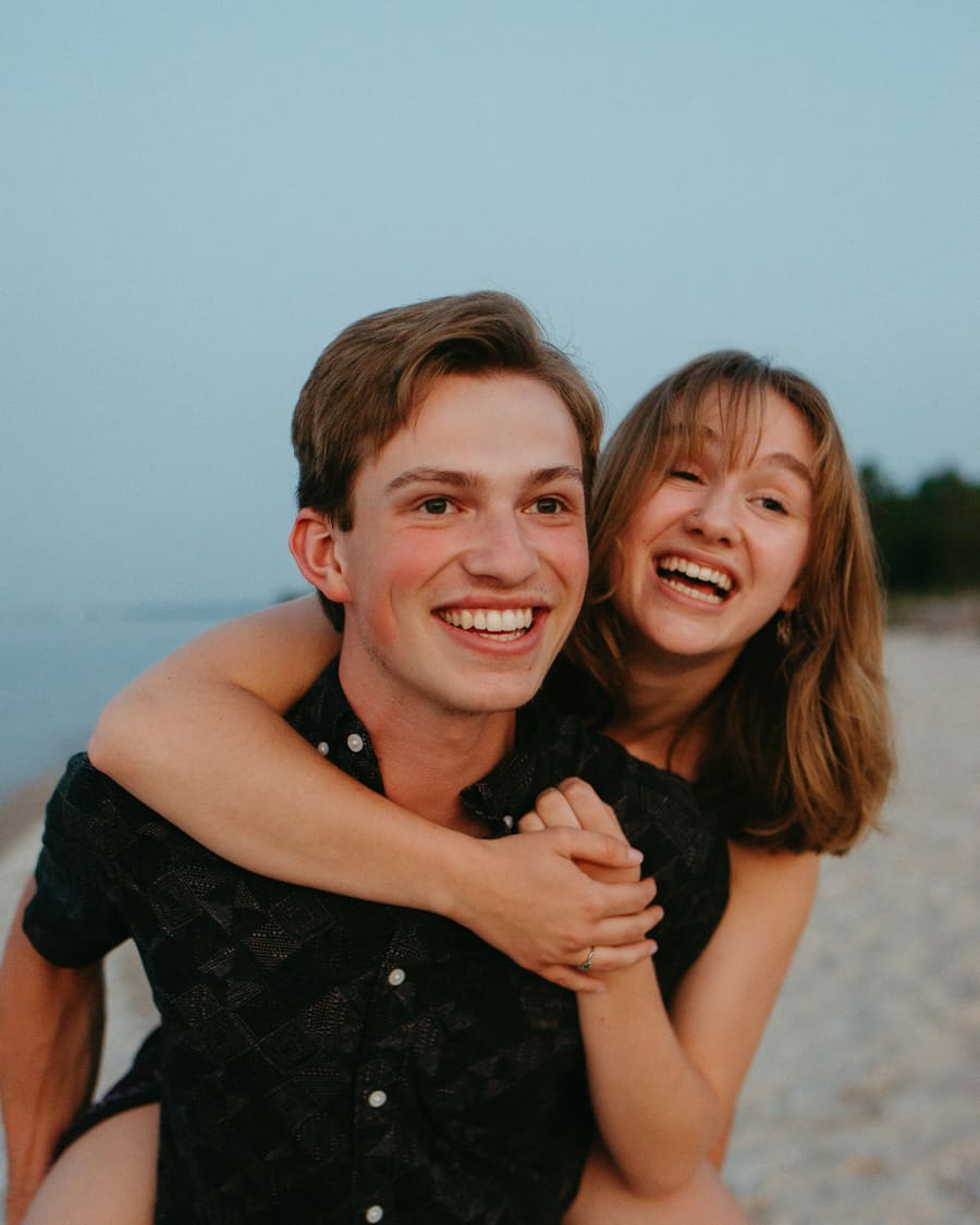 A laughing woman riding piggyback on her companion, a young man in a dark shirt, on a beach under a pale blue sky.