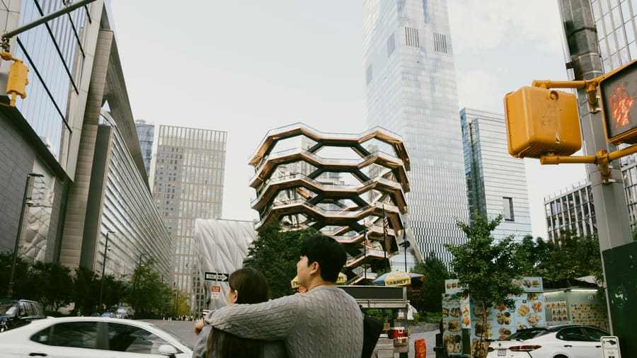 뉴욕 허드슨 야드의 베슬과 유리 빌딩들이 올려다보이는 횡단보도에서 한 여성과 회색 스웨터를 입은 동행자가 뒷모습으로 서 있습니다.