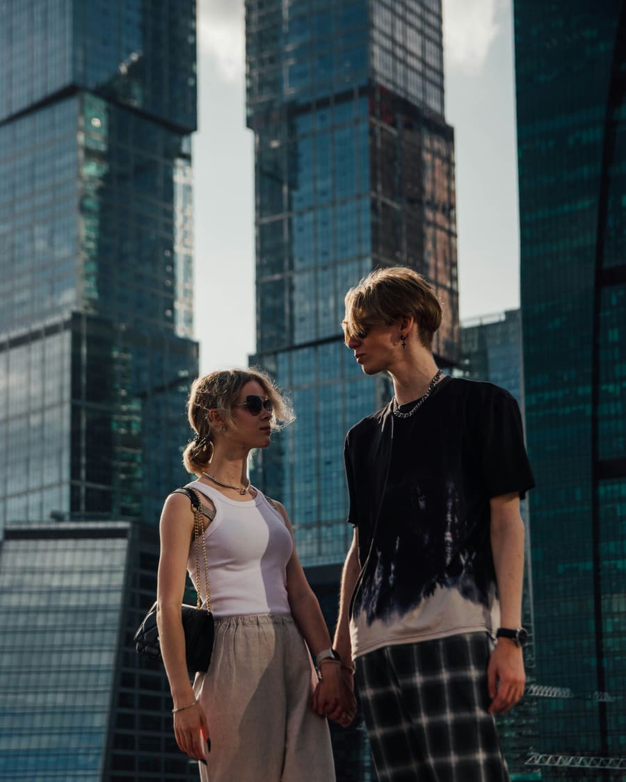 A woman in a white top and sunglasses holding hands with her companion, a man in a black T-shirt, among tall glass towers on a sunny day.