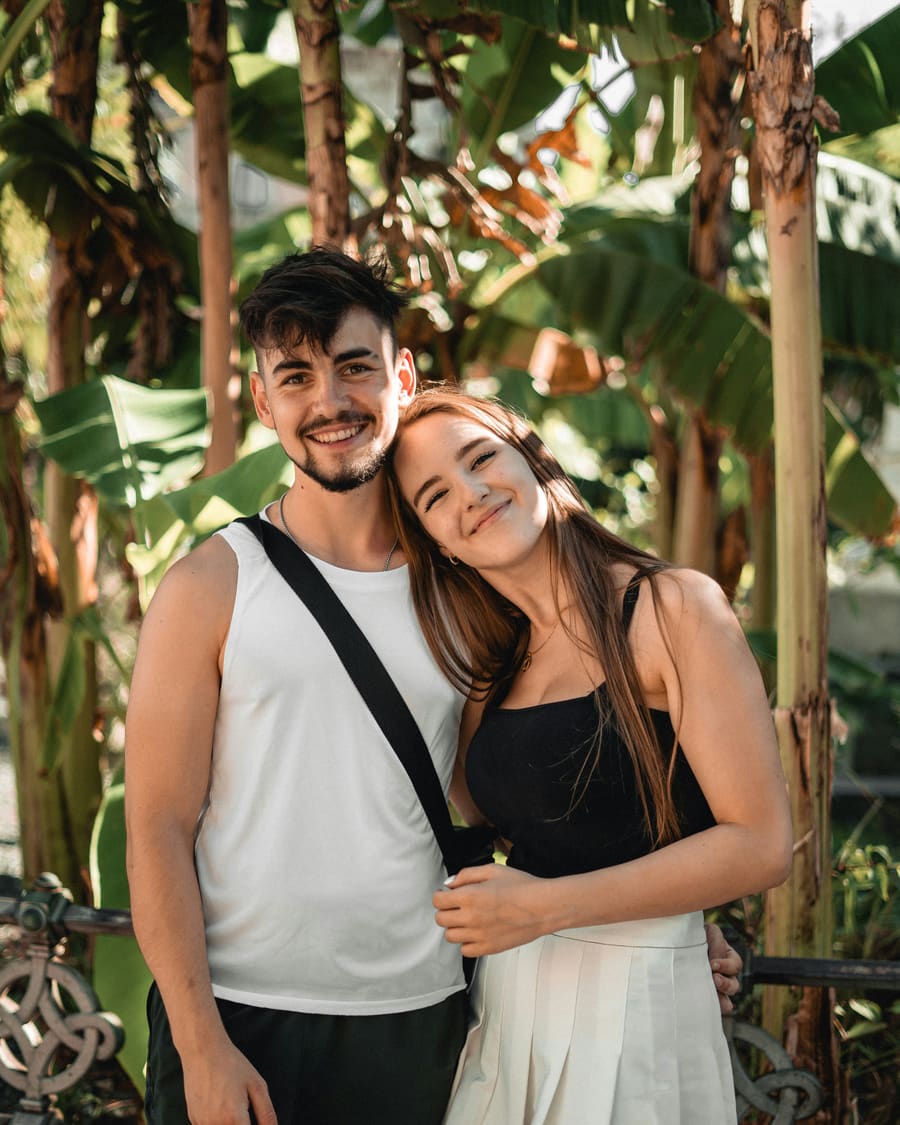 A smiling woman in a black top resting her head on the shoulder of her companion, a man in a white vest, in front of banana plants.